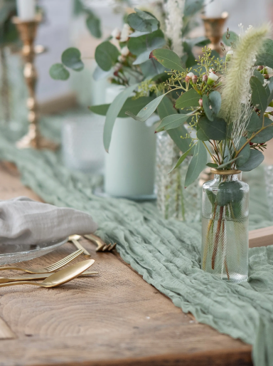 Tischdekoration mit grünen Vasen, Blumen und goldenen Kerzenhaltern auf einem Holztablett.